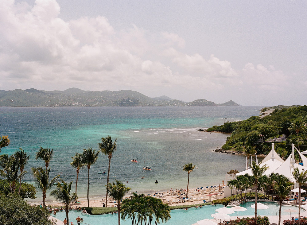 Wedding overlooking a luxurious resort pool in St. Thomas, fringed by swaying palm trees with views of a sun-kissed beach where guests lounge and play, and kayakers paddle in the clear turquoise waters.