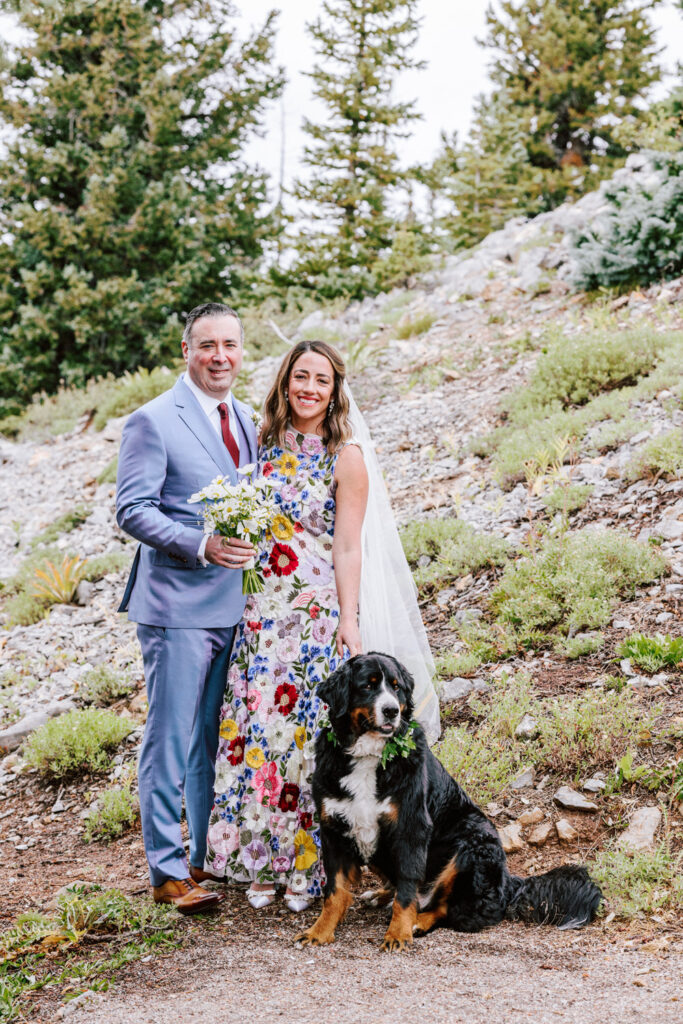 Bride in a floral wedding gown by Rachel Gilbert for her Aspen wedding, next to her groom and their Bernese Mountain Dog, amidst the rustic beauty of Aspen.