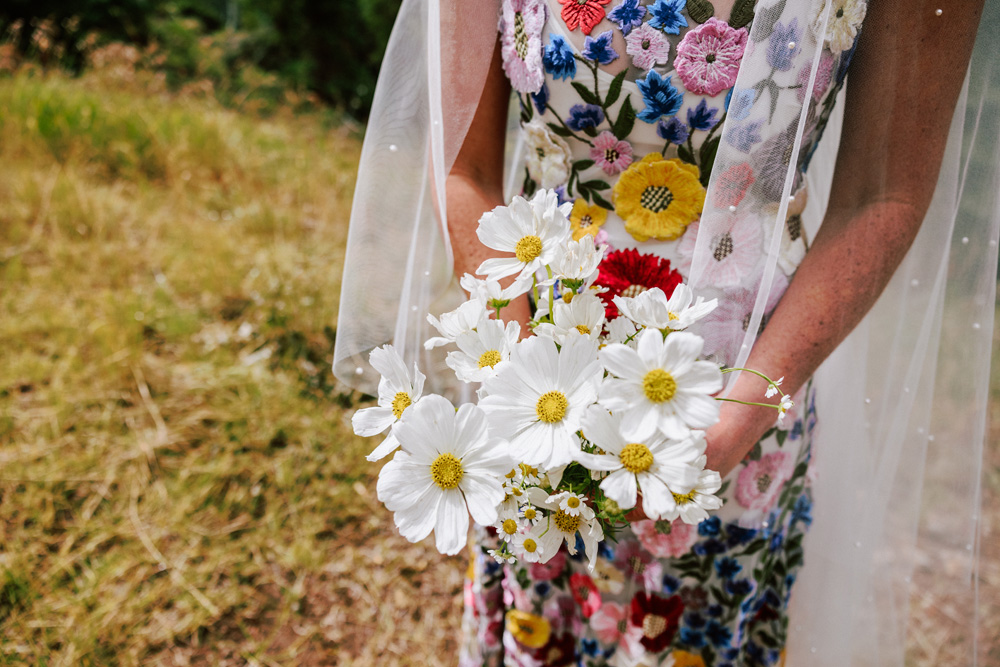 The bride’s bouquet of pristine white daisies offers a sweet, delicate touch against the vivid tapestry of her floral gown by Rachel Gilbert, for her destination wedding in Aspen, Colorado.