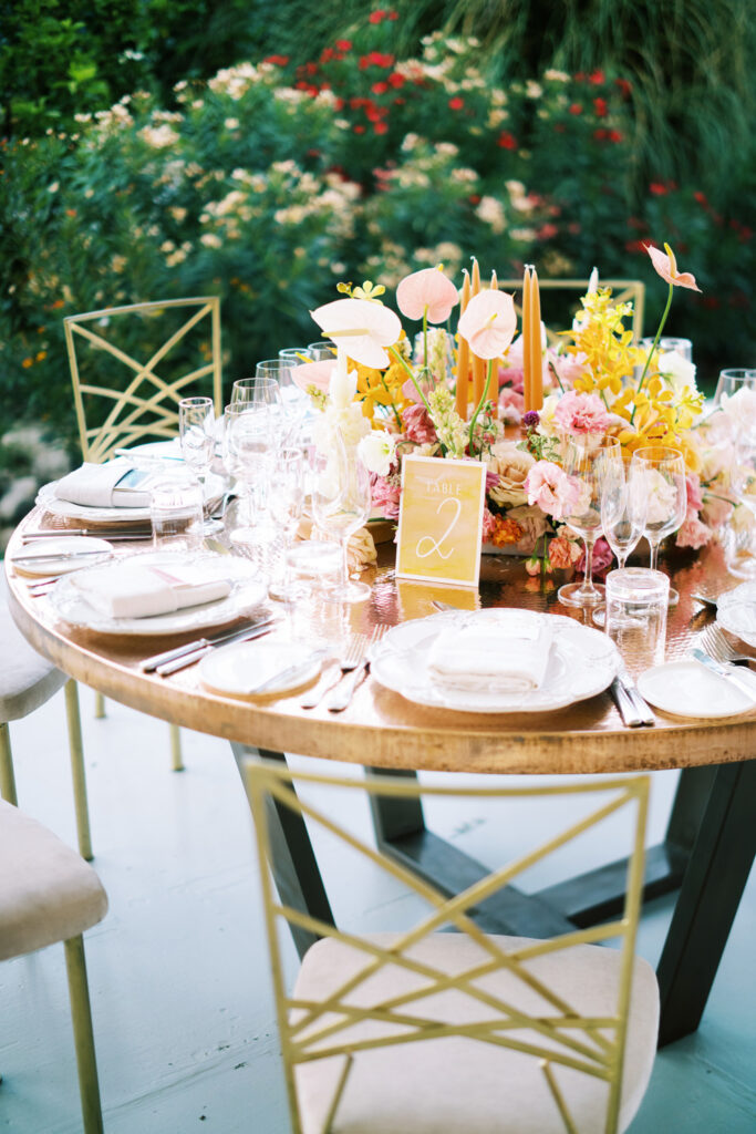 Elegantly set round wedding table with a vibrant floral centerpiece in shades of pink, yellow, and white, complemented by tall candles and a table number sign, with a backdrop of lush red and white flowers, creating a romantic and festive atmosphere for a wedding in San Miguel.