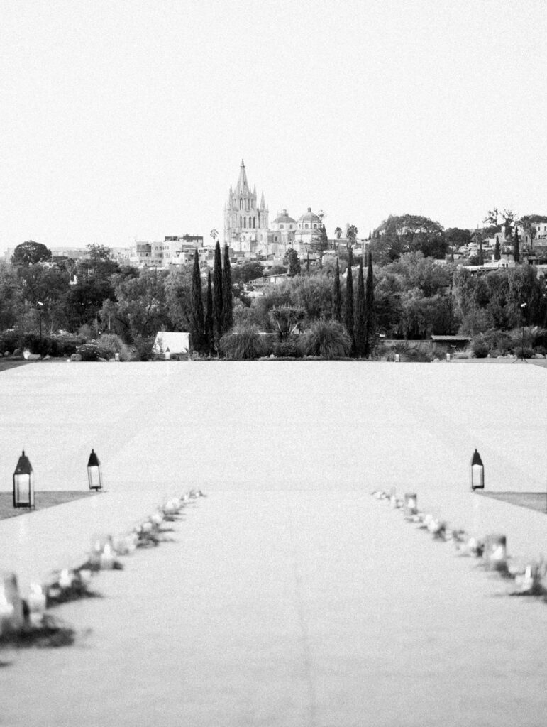 Black and white photograph of a wide ceremonial path lined with lanterns leading towards a distant church and historical buildings in San Miguel Mexico, surrounded by lush greenery and tall cypress trees.