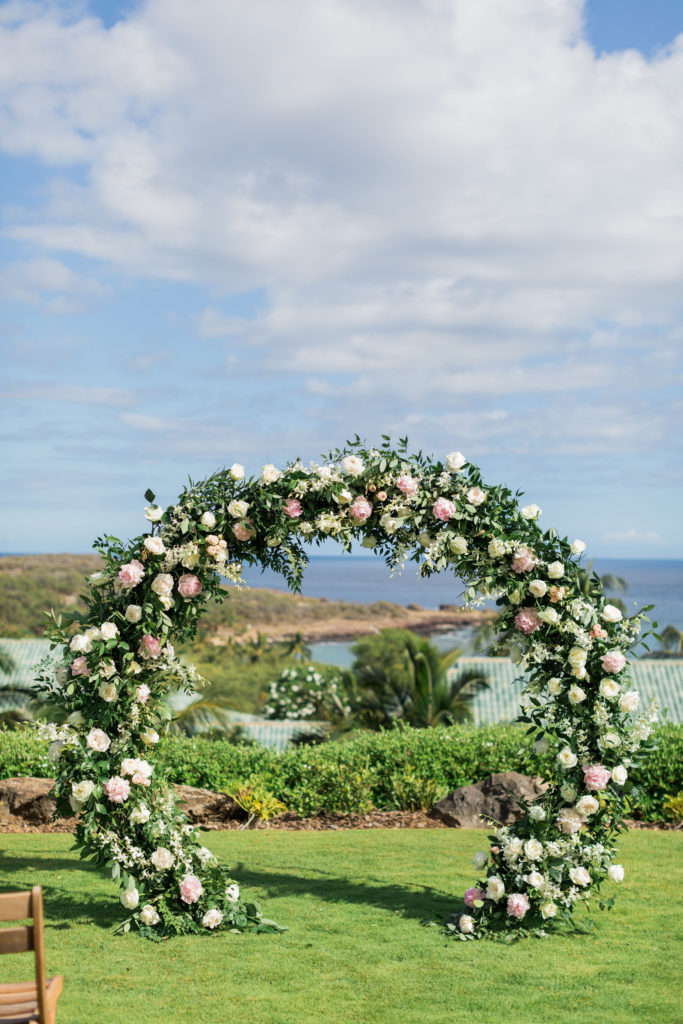 A circular floral arch adorned with pink and white blooms and lush greenery creates a romantic gateway overlooking a serene tropical landscape with palm trees and a calm blue sea.