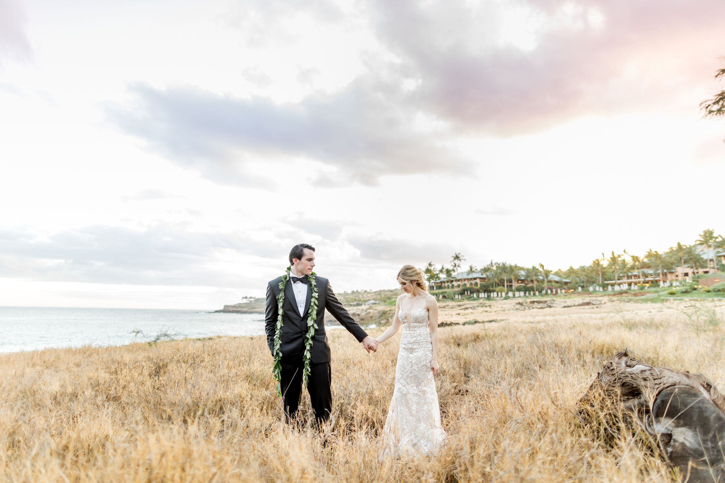 A bride in a lace gown and a groom in a classic tuxedo holding hands and walking through a field of golden tall grass, with the ocean at dusk and a distant beach beneath a pastel sky in the background.