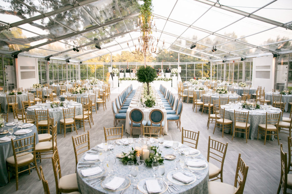 An elegant wedding reception setup inside a clear tent with transparent walls and ceiling, showcasing arranged tables with linen and floral centerpieces, and a live band set up at the far end.