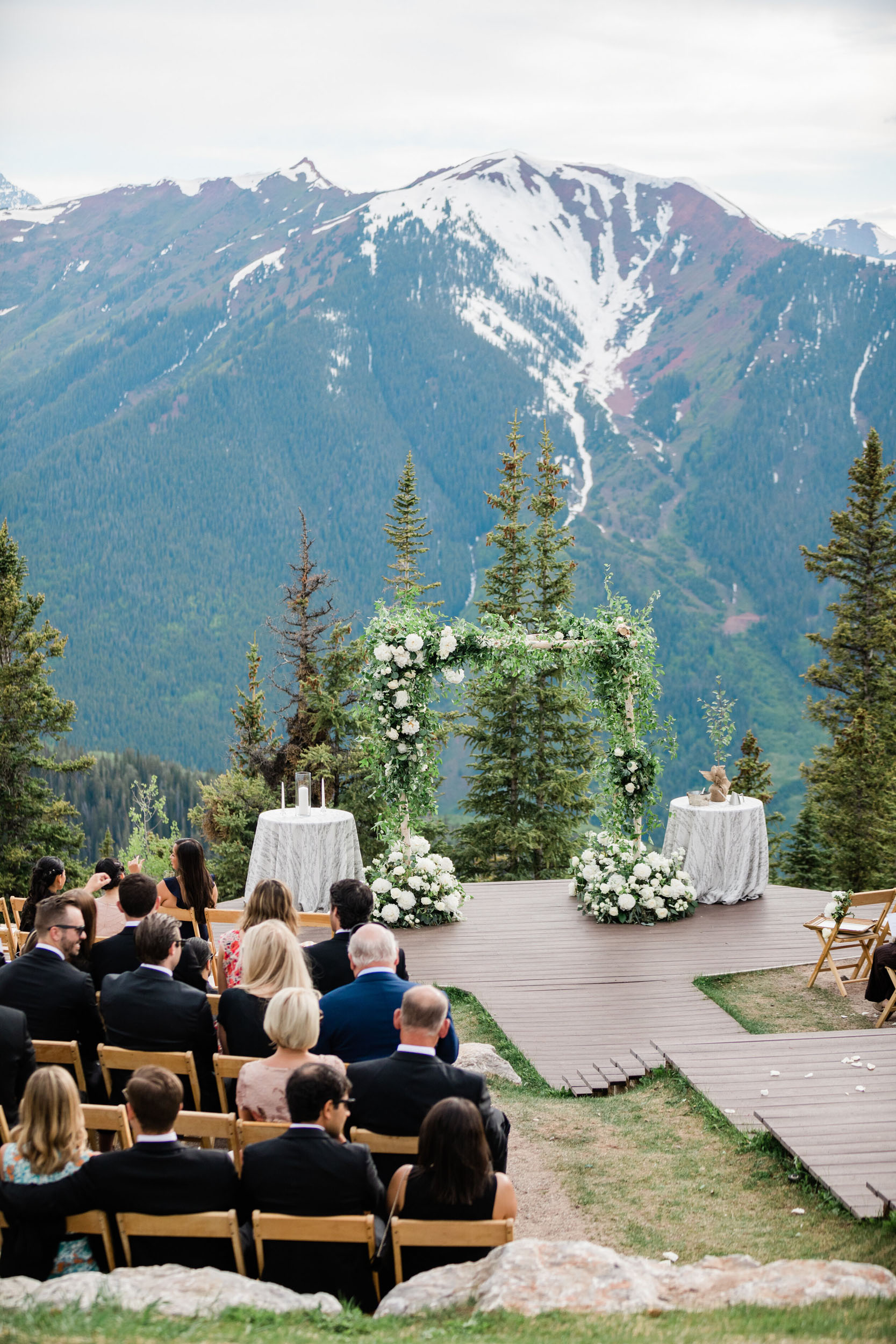 Guests seated on wooden chairs at an outdoor wedding ceremony with a floral arch, facing snow-capped mountains in the background.
