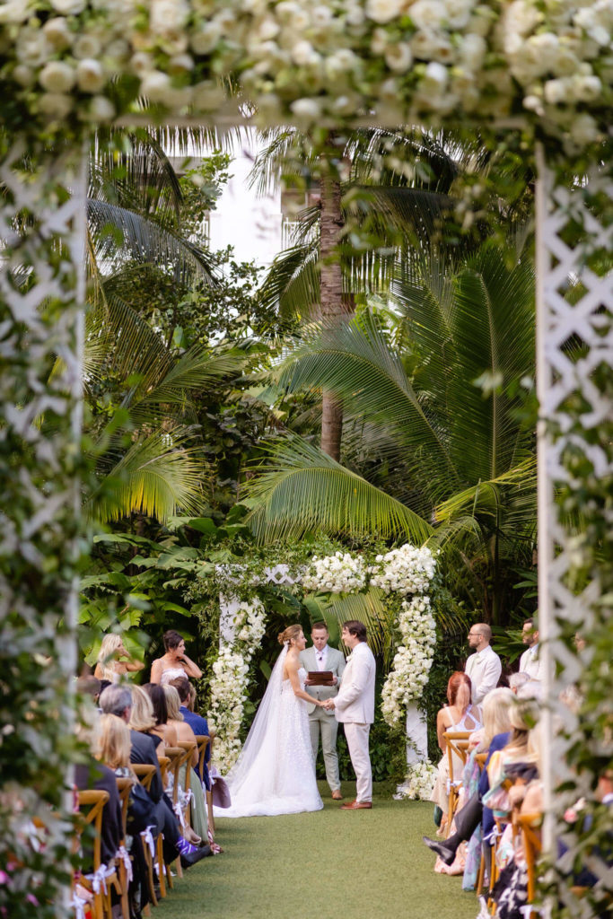 A bride in a white lace gown and a groom in a white suit stand at the altar, surrounded by lush tropical greenery and white floral decorations, as guests watch the intimate ceremony.