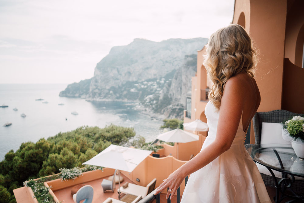 A bride in a flowing white gown gazes from a terracotta balcony over the azure waters of the Capri coast, with cliffs and scattered boats in the tranquil sea below.