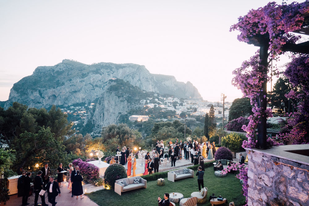 Elegantly dressed guests at a wedding cocktail hour on a verdant terrace, with the iconic Faraglioni rocks in the backdrop, as evening falls on the stunning landscape of Capri.