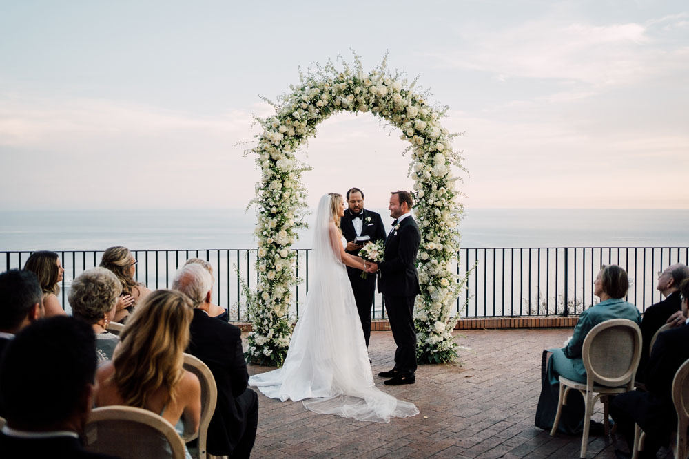 The bride and groom hold hands at their wedding ceremony under a white floral arch, with guests seated around them, against the panoramic backdrop of the Italian coast.