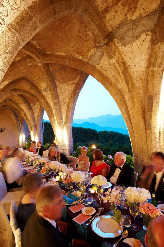 Guests dine elegantly at a long table set within open Italian stone arches, enjoying the ambient lighting and the breathtaking mountain views beyond.