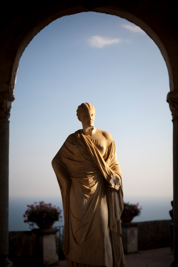 A classical statue stands in the foreground of an archway, basking in the soft glow of sunset at the historic Hotel Villa Cimbrone, with the sea horizon in the distance.