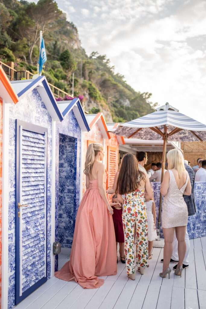 Women in elegant summer attire congregate beside vibrant, patterned beach huts with the picturesque cliffs of the Amalfi Coast rising in the background.