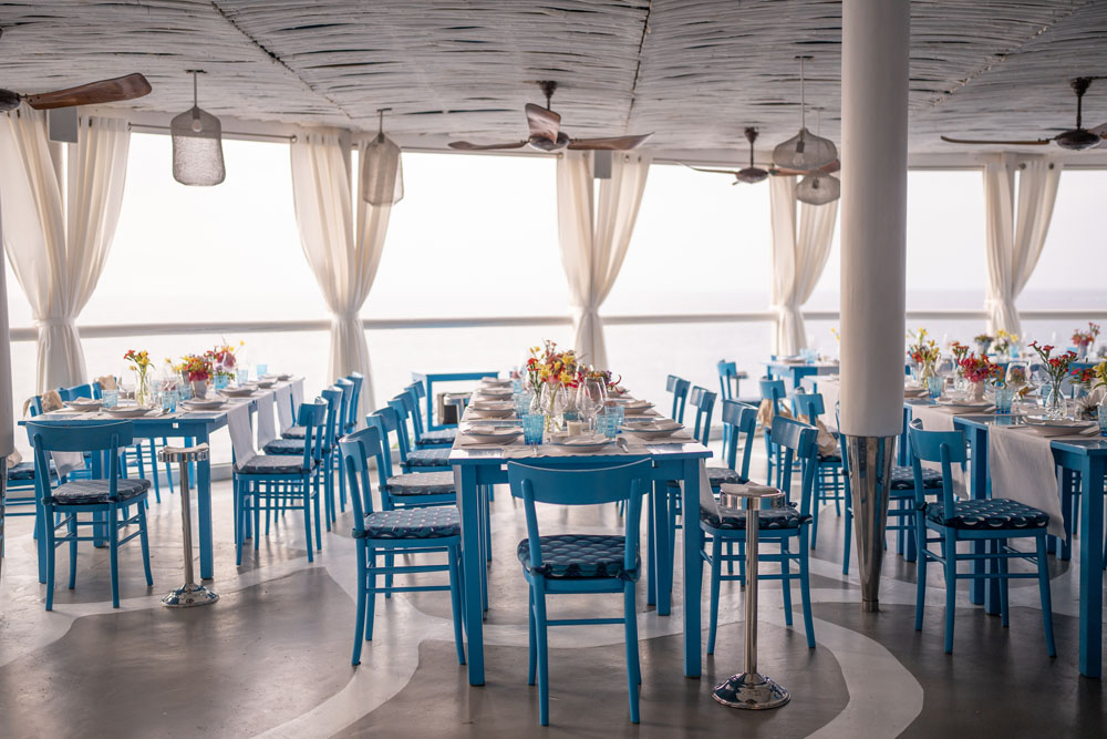 Elegantly set tables with bright blue chairs arranged in a chic, nautical-themed seaside dining area, complete with breezy curtains and a view of the serene ocean.