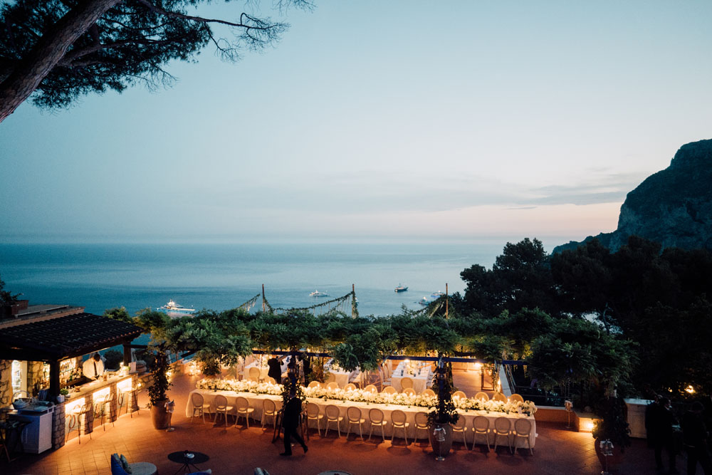 A twilight view of an outdoor wedding reception on a terrace by the sea, with long tables under a canopy of floating greenery, and the calm ocean dotted with boats in the background.