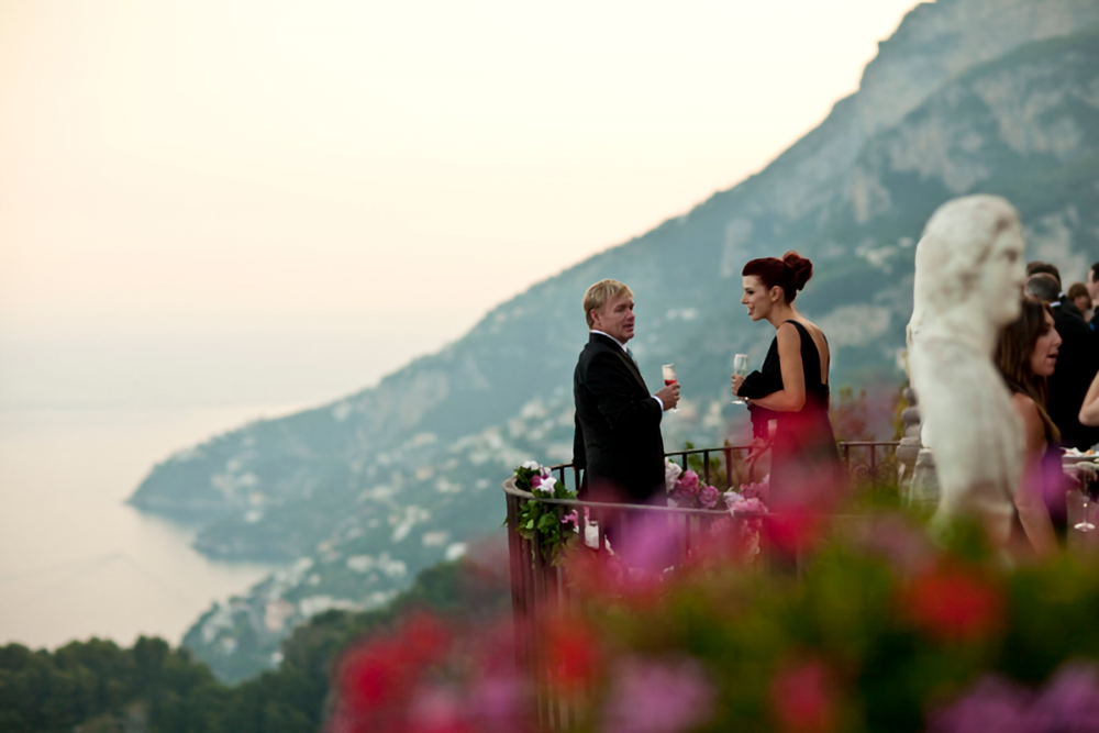 Wedding guests in formal attire engaged in conversation, holding wine glasses on a balcony with a soft-focus background of majestic cliffs and the tranquil sea at sunset.