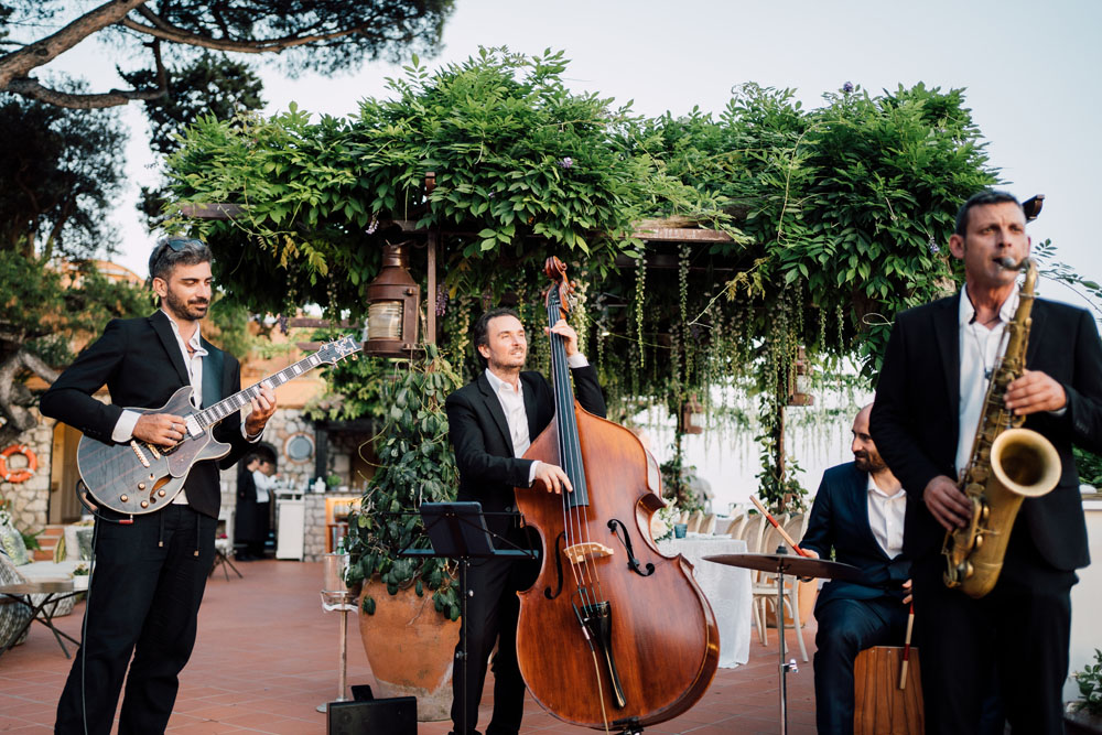 A live jazz band consisting of a guitarist, cellist, and saxophonist playing soulful music at a wedding venue adorned with greenery and Mediterranean-style decor.