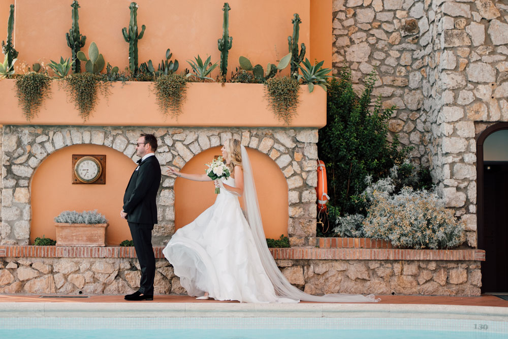 Bride in a flowing white gown reaching out to tap the shoulder of the groom for a 'first look' beside a pool, with a backdrop of traditional Mediterranean architecture and succulent plants.