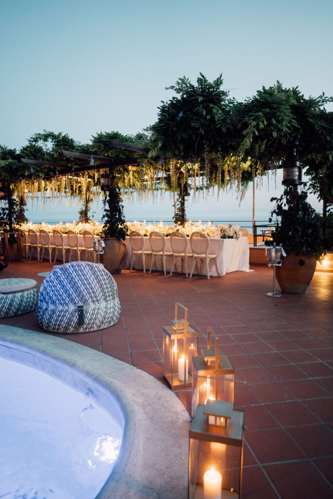 Lit lanterns line the edge of a pool on a terracotta-tiled terrace, leading to an elegantly set wedding reception area under a canopy of greenery with a view of the tranquil sea at dusk.