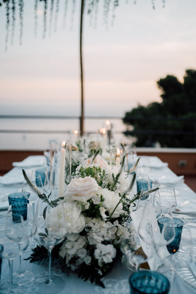 An intimate wedding table setting featuring a white floral centerpiece, elegant glassware, and fine tableware on white linen, illuminated by the soft glow of candlelight.