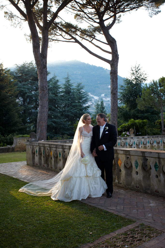 A bride and groom share a tender moment amidst the lush gardens of Il Riccio Beach Club, with stone balustrades and the serene backdrop of the Amalfi hills framing their embrace.