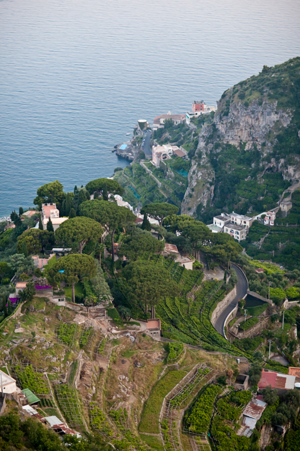 An aerial view captures the dramatic and lush cliffsides of the Amalfi Coast, with terraced gardens and winding roads leading down to the serene Mediterranean Sea, seen from the vantage point of Il Riccio Beach Club.