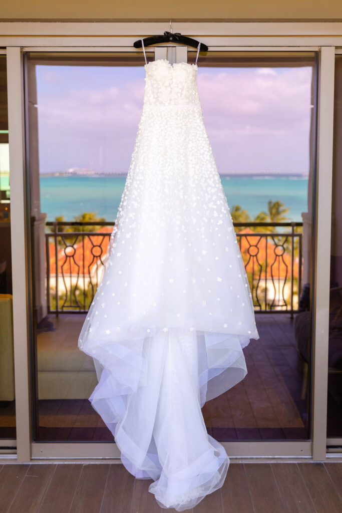 A bride's exquisite wedding gown hangs in anticipation in front of a sliding glass door, reflecting the vibrant hues of the ocean and the skyline of the Bahamas.