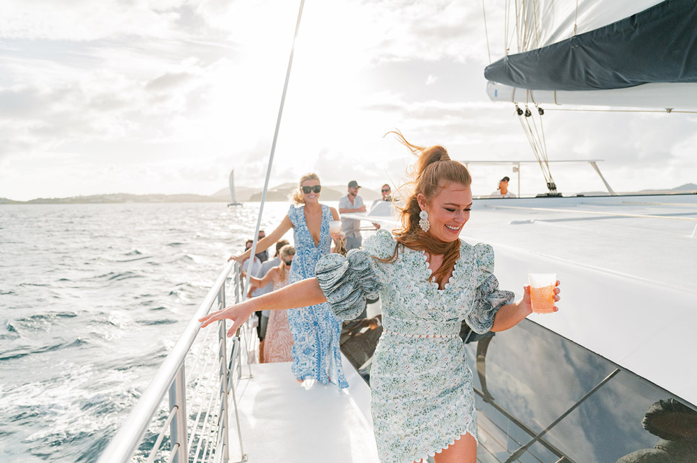 A woman with a joyful smile holds a drink on the sunlit deck of a sailboat, cruising through the refreshing ocean breeze.