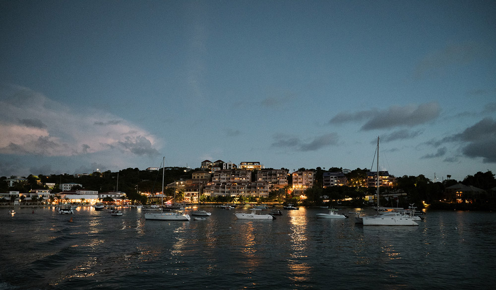 Twilight descends on a tranquil harbor in St. Thomas, where boats gently rock in the water, and the lights of a resort twinkle against the evening sky.