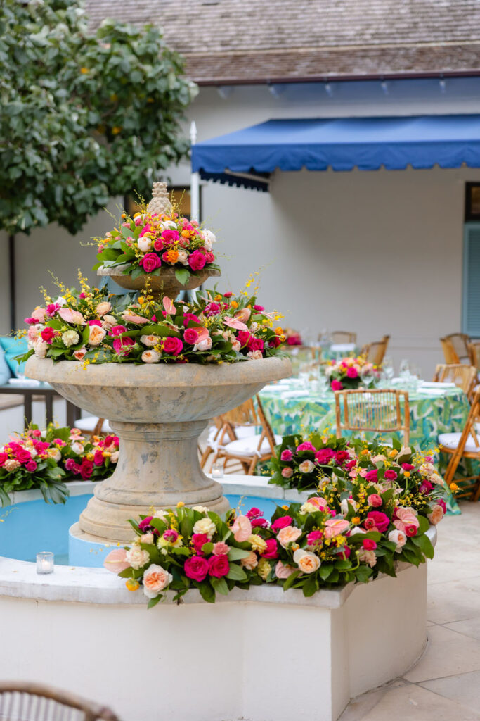 An ornate stone fountain is richly adorned with vibrant pink and peach roses, creating a stunning floral display in a charming courtyard setting.