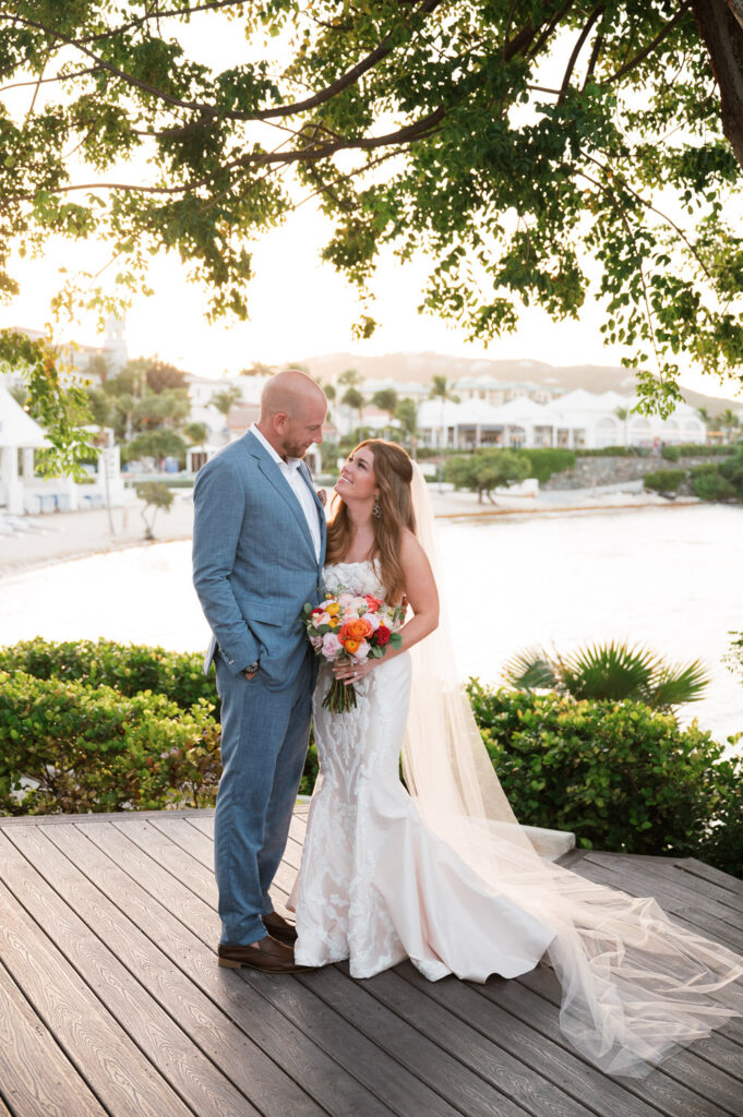 A bride in a lace wedding gown holding a vibrant bouquet lovingly gazes at the groom on a wooden deck, with the tranquil waters and a picturesque St. Thomas resort in the background.