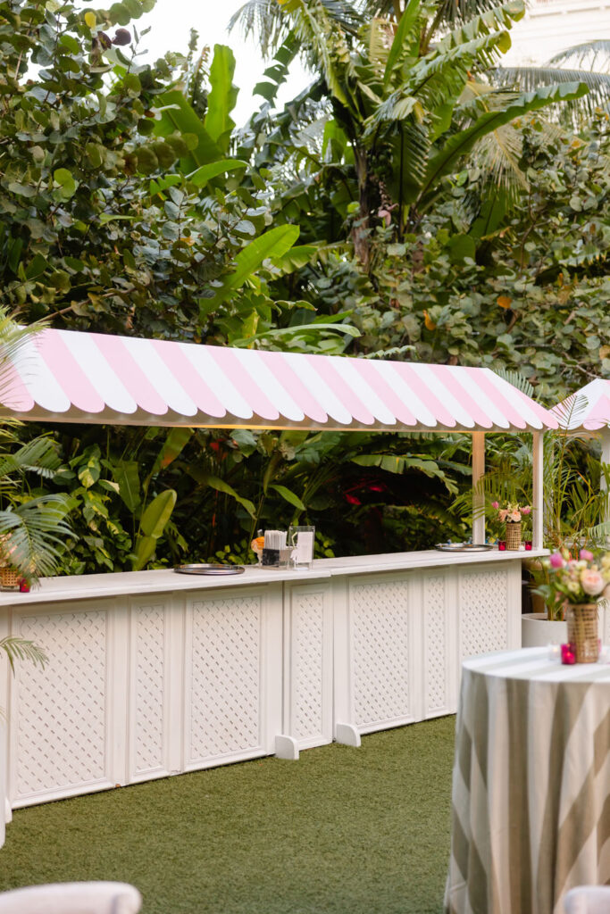 A chic wedding bar setup under a striped pink and white awning stands ready for guests, surrounded by lush tropical foliage that enhances the festive atmosphere.