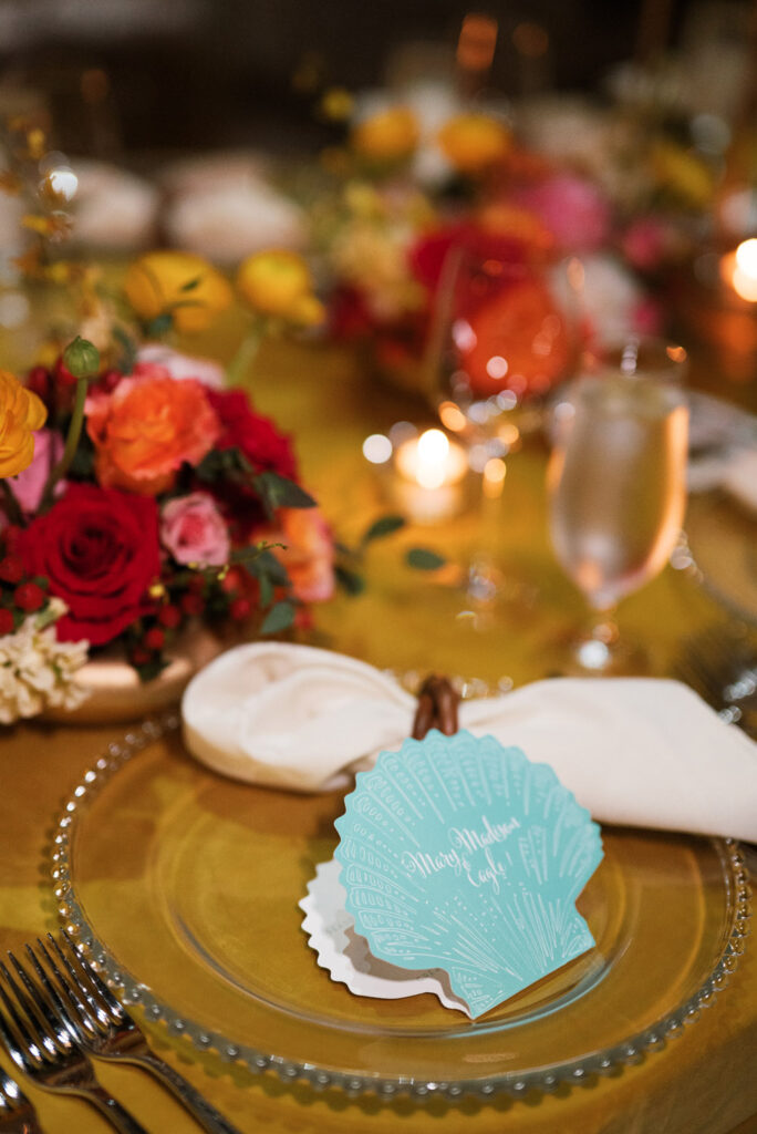 An intimate close-up of a wedding table setting, featuring a clear charger and a personalized seashell place card, with a backdrop of colorful flowers and warm candlelight.