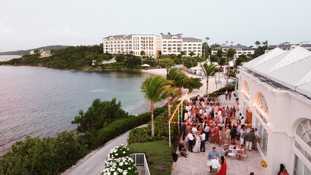 A bustling gathering of people outside a white building, enjoying the evening under swaying palm trees, with the water reflecting the pink hues of dusk.