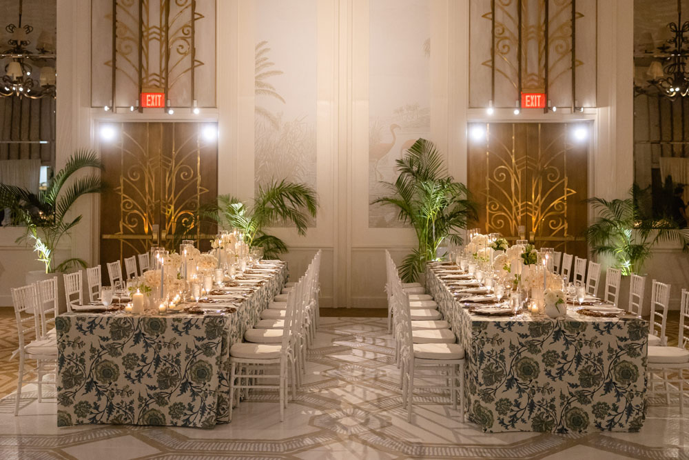 An opulent wedding reception setup at The Grand Salon in Rosewood Baha Mar, featuring tables draped with floral-patterned linens, elegant white chairs, and ambient candlelight, complemented by lush palm decorations.