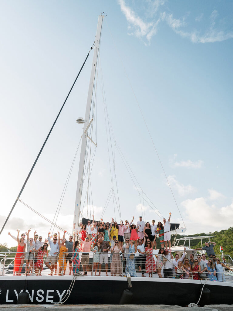 A joyful group of people posing for a photo aboard the deck of a sailboat, with clear skies above and the excitement of the open sea around them.