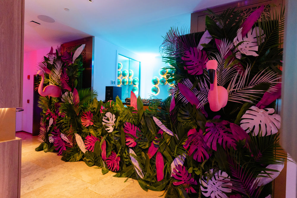 A vibrant DJ booth at a wedding, creatively adorned with pink, white, and green palm leaves, featuring two whimsical lawn flamingos on each side, under the glow of neon lights.