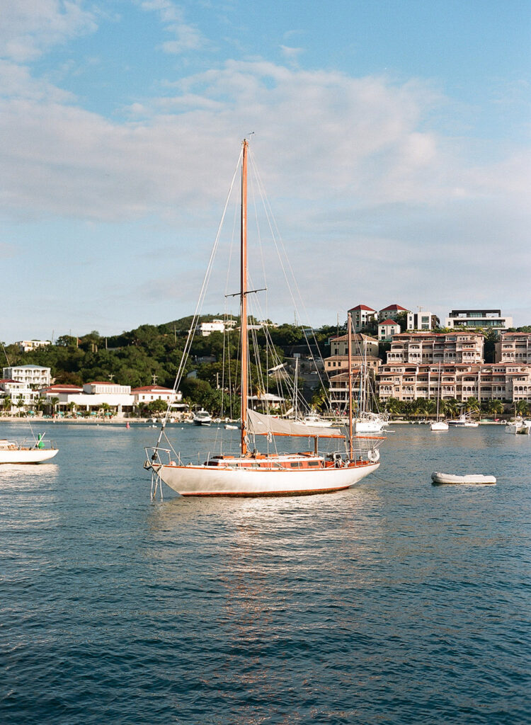 A classic sailboat floats gracefully in the serene blue waters of a harbor, accompanied by other boats, with a backdrop of coastal buildings under a soft sky.