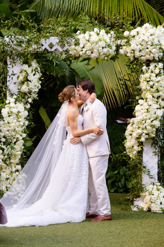 The newlyweds share a kiss at the conclusion of their wedding ceremony, standing under a beautiful white floral arch that frames this romantic moment, surrounded by lush greenery.
