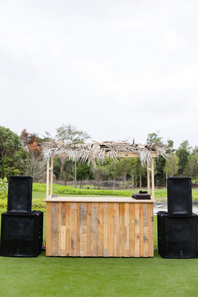 A rustic wooden cabana-style DJ booth is set up on a neatly trimmed lawn for a wedding, flanked by two large speakers, ready to fill the space with music.