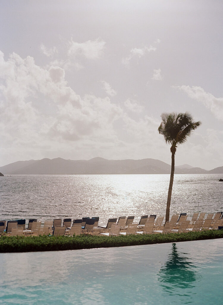 Scenic view from the Ritz-Carlton in St. Thomas, showcasing the tranquil infinity pool leading to a sunlit beach adorned with lounge chairs and a single palm tree against a backdrop of distant hills.