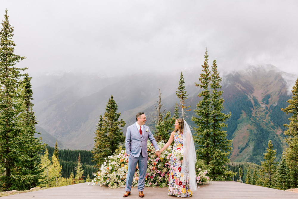 Bride and groom holding hands on a wooden deck, exchanging loving glances. The bride stands out in a unique floral wedding dress, next to a lavish floral arrangement, with the majestic Rocky Mountains serving as a breathtaking backdrop.