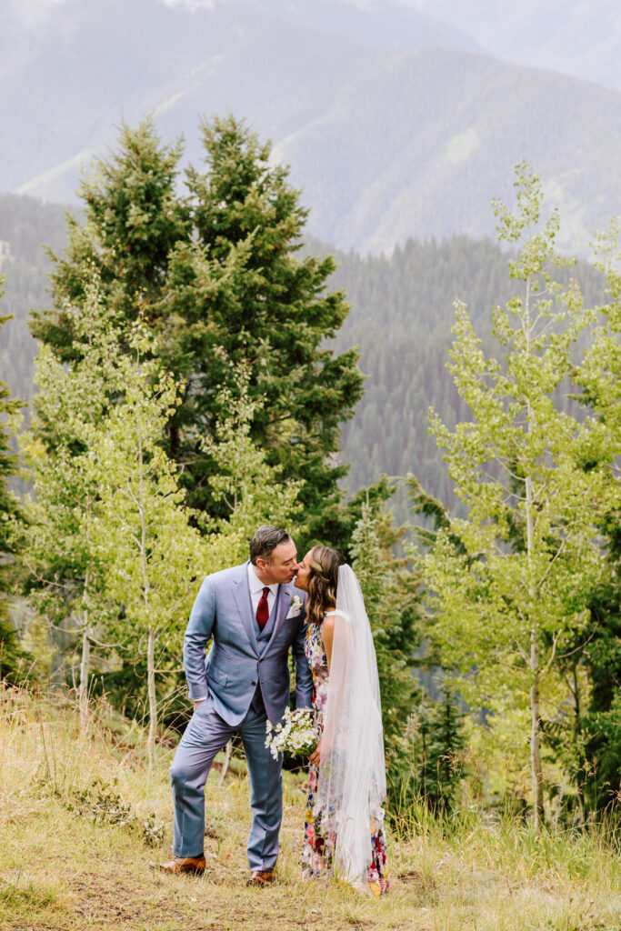 Bride and groom share a tender kiss amidst the natural splendor of the Rockies, with lush evergreens framing the intimate moment. The bride’s floral gown adds a splash of color to the verdant surroundings.