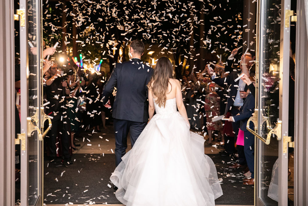 The bride and groom make their grand exit through a shower of confetti, with guests cheering, captured at the moment they step out of the venue into a night of celebration.