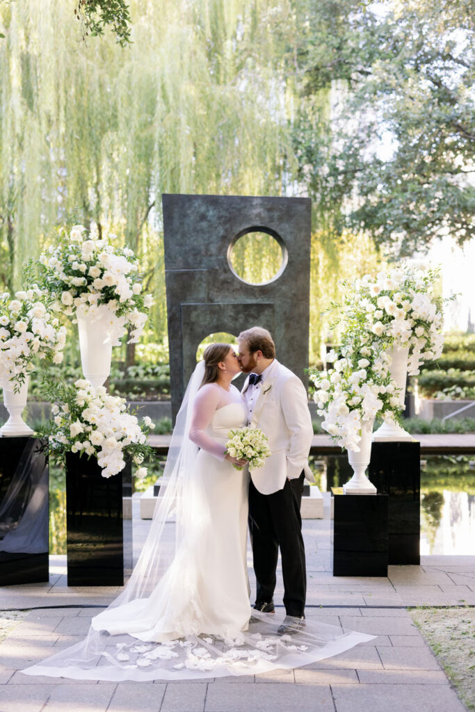 A bride and groom share a kiss at an outdoor altar under a willow tree, flanked by tall white floral arrangements on pedestals, symbolizing a romantic wedding ceremony.