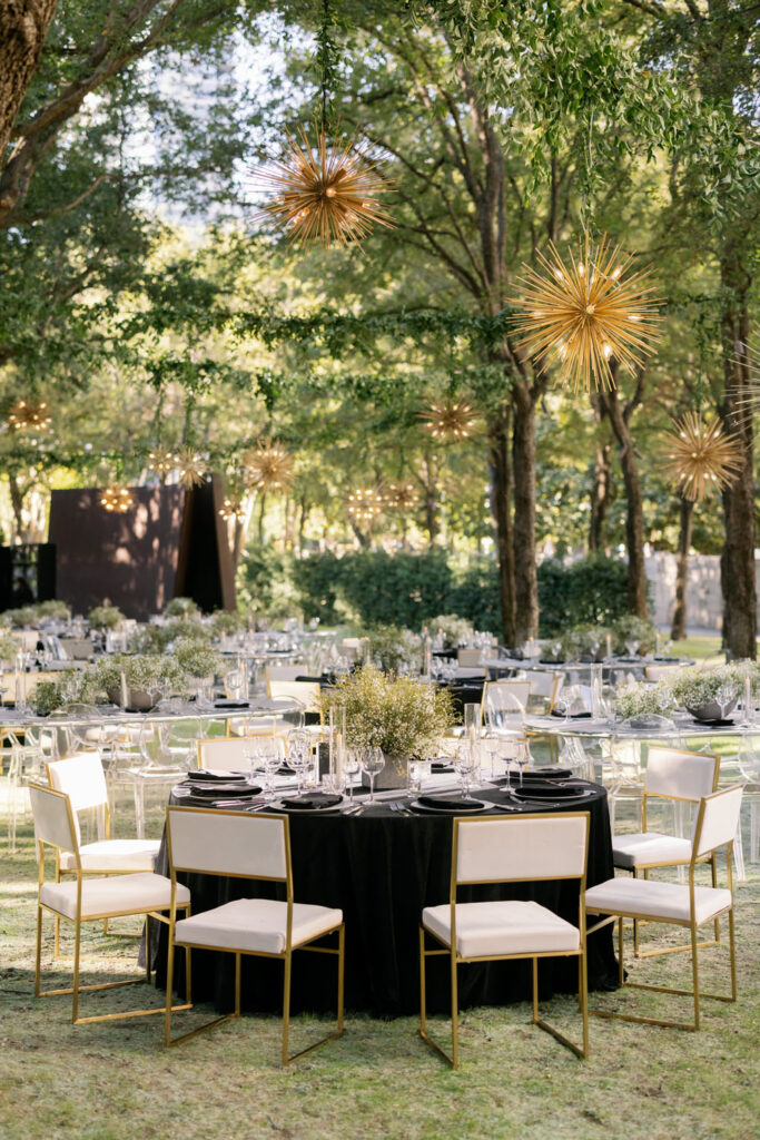 Outdoor wedding reception area with chairs and black tablecloths, adorned with golden starburst light fixtures hanging from the trees, creating a whimsical evening atmosphere.