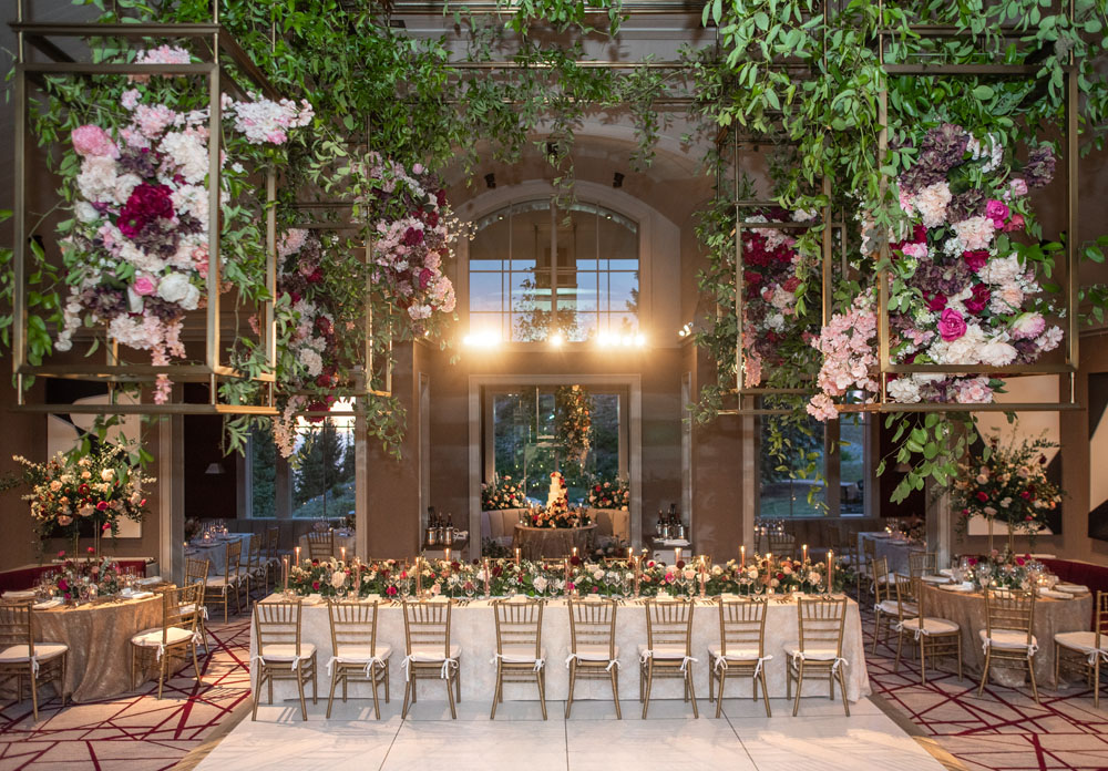 Elegant wedding reception in a grand hall with lush hanging floral installations in golden geometric frames, and tables set beneath with chic decor and soft lighting.
