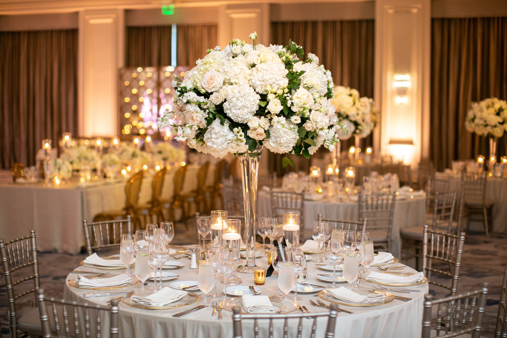 Sophisticated wedding reception table featuring a tall, opulent white floral centerpiece, surrounded by silver chiavari chairs and glowing candlelight, set in a luxurious ballroom.