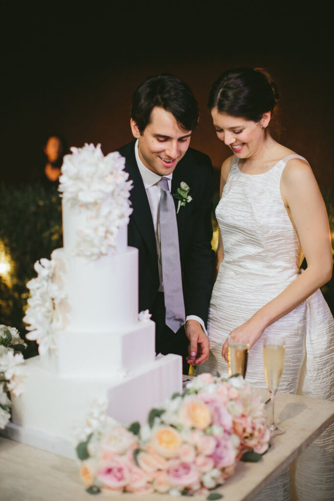 Bride and groom at their wedding, smiling as they cut into a multi-tiered white cake, with lush floral arrangements and champagne glasses on the table.