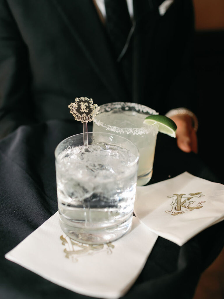 A close-up of elegant wedding drinks, one with a monogrammed stirrer, on a napkin with the couple’s initials, hinting at the meticulous details of the celebration.