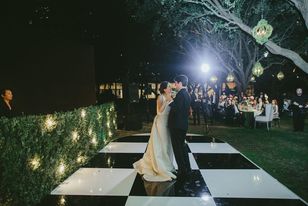Newlyweds sharing their first dance on a checkered dance floor outdoors at night, surrounded by the warm glow of lights and guests seated at tables in the background.
